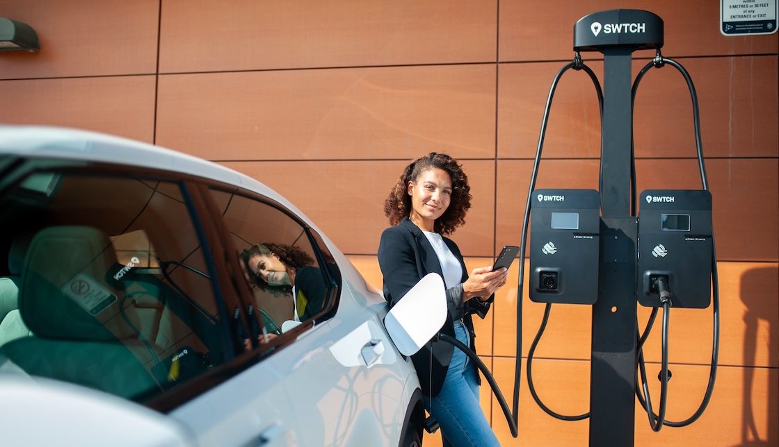 A woman standing next to an EV plugged into a charger
